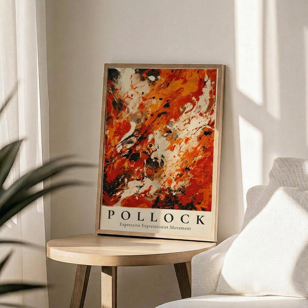 May include: Abstract expressionist artwork poster with vibrant orange, red, white and black paint splatters and brushstrokes, displayed in light wood frame on round wooden side table next to beige upholstered chair. Natural sunlight creates soft shadows on white wall behind. Green plant leaves visible in corner of minimalist, modern living room setting.