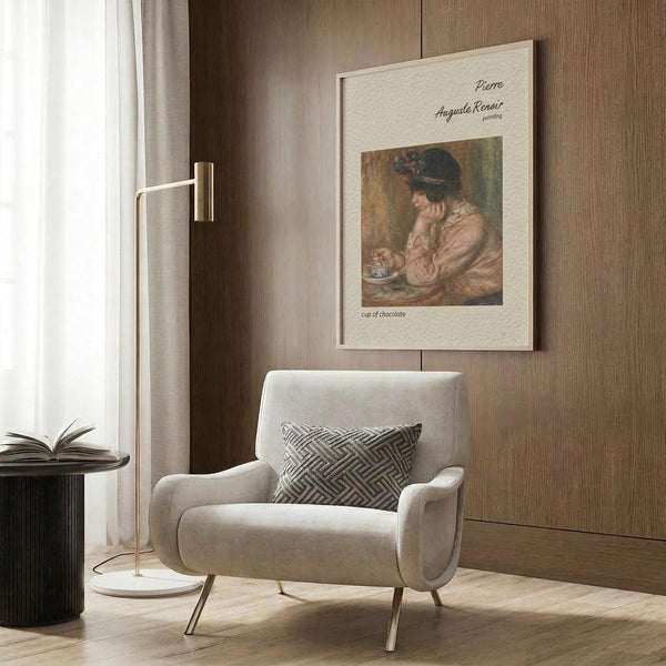 May include: Framed art print of Pierre-Auguste Renoir's "Cup of Chocolate" painting showing a woman in pink dress with dark hat holding a teacup, displayed on warm wood-paneled wall above beige upholstered armchair with geometric patterned pillow, alongside brass floor lamp and dark round side table with open book near window with white curtains