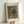 May include: Hand holding a framed art print showing an impressionist still life painting of wildflowers in an earthenware vase. The artwork features loose brushstrokes in muted greens, blues, reds and yellows against a gray background. White matting displays the title "FLOWERS IN A VASE" at top and "PIERRE AUGUSTE RENOIR 1866" at bottom. Light wood frame against white wall.