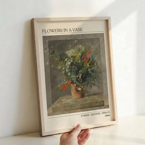 May include: Hand holding a framed art print showing an impressionist still life painting of wildflowers in an earthenware vase. The artwork features loose brushstrokes in muted greens, blues, reds and yellows against a gray background. White matting displays the title "FLOWERS IN A VASE" at top and "PIERRE AUGUSTE RENOIR 1866" at bottom. Light wood frame against white wall.