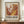 May include: Framed exhibition poster featuring abstract paint swirls in warm oranges, browns, yellows and white with scattered blue dots, displayed on wooden credenza beside white ceramic vase and stacked books against cream paneled wall