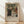 May include: Framed exhibition poster featuring a classical family portrait painting with adults and children in period dress, displayed against white wall on wooden floor. Poster shows "PIERRE AUGUSTE exhibition" text below artwork dated 1896, titled "The Artist's Family." Natural wood frame with beige matting, soft window light creating diagonal shadows across the scene.