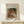 May include: Framed art print of Renoir's "Child with Toys" painting showing woman in rust-colored dress with blonde child playing with small figurines, displayed on light wood console table next to white wrapped gift with evergreen sprig, against cream wall with natural lighting