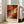 May include: Framed abstract art print with vibrant orange, red, white and black swirling paint strokes in expressionist style, displayed on light wood floor against white wall next to beige upholstered chair. Natural sunlight streams through tall windows creating soft shadows. The artwork features dynamic fluid movements and bold color contrasts in a light wood frame.