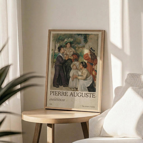 May include: Framed exhibition poster displaying Renoir's "The Artist's Family" painting from 1896, showing a family group with adults and children in period dress against a garden backdrop. The poster sits on a round wooden side table next to a beige upholstered chair, with soft natural lighting and plant shadows creating an elegant home gallery setting.