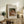 May include: Framed reproduction of Renoir's "Child with Toys" showing a woman in coral dress holding a blonde child with small toy figures, displayed on rustic wooden sideboard alongside fern plant, ceramic vase, and books in warm neutral living room setting.