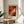 May include: Abstract expressionist art poster with vibrant orange, red, black and white paint swirls displayed in thin wooden frame on round wooden side table. Neutral beige armchair and green plant leaves visible in softly lit modern living room with white walls and natural window light creating gentle shadows.