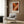 May include: Abstract expressionist artwork poster with vibrant orange, red, black and white paint splatters in white frame, displayed above cream upholstered armchair with geometric patterned pillow, next to brass floor lamp and dark round side table with open book, against wood-paneled wall in modern living room setting.