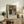 May include: Framed exhibition poster featuring Renoir's family portrait painting displayed on rustic wooden sideboard alongside fern plant, ceramic vase, and stacked books. Poster shows adults and children in period clothing with "PIERRE AUGUSTE exhibition" text below. Warm neutral tones throughout cozy living room setting with upholstered chair visible.
