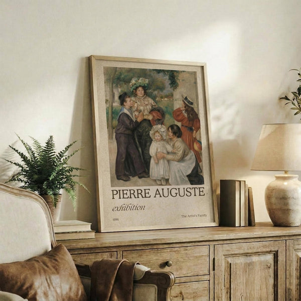 May include: Framed exhibition poster featuring Renoir's family portrait painting displayed on rustic wooden sideboard alongside fern plant, ceramic vase, and stacked books. Poster shows adults and children in period clothing with "PIERRE AUGUSTE exhibition" text below. Warm neutral tones throughout cozy living room setting with upholstered chair visible.
