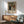 May include: Framed exhibition poster featuring a classical painting of a family group with adults and children in period dress, displayed above a wooden sideboard with white ceramic lamp. Mint green mid-century modern armchair with wooden arms sits nearby in bright, naturally lit room with white walls and hardwood floors.