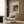 May include: Framed Renoir reproduction poster showing woman in brown dress with blonde child holding toys, displayed on wood paneled wall above beige upholstered armchair with geometric patterned pillow. Modern reading corner with brass floor lamp and dark side table with open book. Warm neutral tones throughout contemporary interior setting.