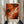 May include: Person holding framed abstract expressionist artwork with vibrant orange, red, white and black swirling paint strokes in light wood frame. Dried pampas grass visible to left side against neutral curtained background. Hands wearing dark pinstriped sleeves display the energetic, fluid composition featuring bold gestural brushwork and dynamic color movement.