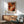 May include: Abstract expressionist art poster with vibrant orange, red, and black paint swirls in wooden frame, displayed above mid-century modern wooden credenza with white ceramic lamp. Sage green upholstered chair with wooden arms sits nearby on hardwood floor. Soft natural light creates shadows on white wall in minimalist living room setting.