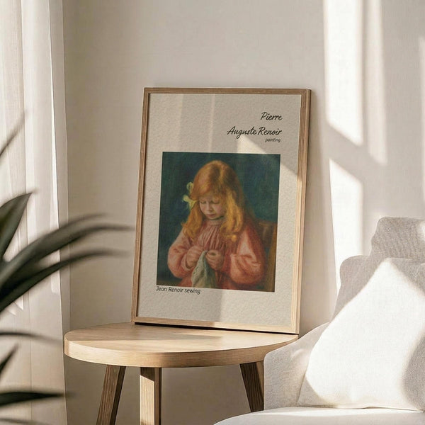 May include: Framed poster print displayed on round wooden side table next to beige upholstered chair. Print shows young girl with reddish hair in pink dress sewing, with text identifying it as Pierre Auguste Renoir painting "Jean Renoir sewing". Warm sunlight streams across neutral-toned room with plant shadows visible on white wall.