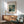 May include: Framed art print of Renoir's impressionist painting showing two figures dining at a restaurant, displayed on white wall above wooden sideboard with table lamp. Mid-century modern green upholstered chair with wooden frame in foreground, natural sunlight creating shadows across the minimalist room setting.