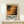 May include: Framed abstract art poster with chaotic paint splatters in warm oranges, browns, and blacks displayed on light wood shelf against cream wall. Small white gift box with twine sits beside the natural wood frame. The artwork features dynamic brushstrokes and paint drips in an expressive style with exhibition text below the image.