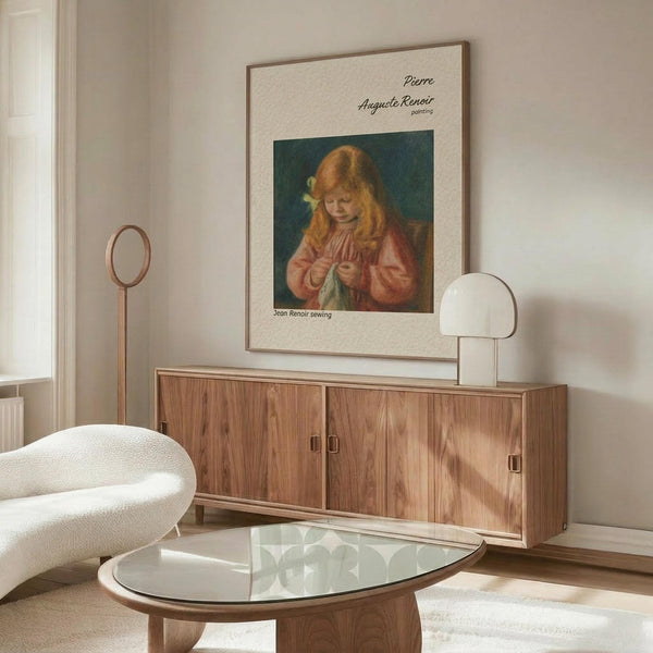 May include: Framed vintage art print of young girl with red hair sewing, displayed on wooden credenza in modern living room with cream chair, glass coffee table, and white table lamp. Print shows soft impressionist painting style with warm tones against neutral wall.