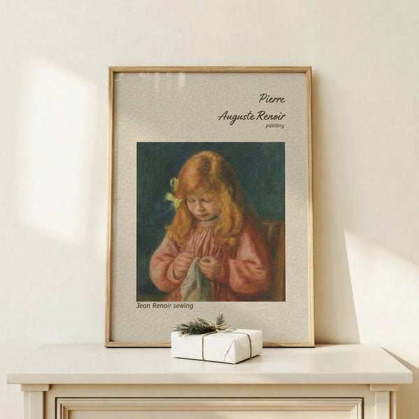 May include: Framed art print of Renoir painting showing young girl with reddish hair in pink dress sewing, displayed on light wood console table next to small wrapped gift with greenery. Warm beige wall background, natural lighting creating soft shadows.