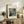 May include: Framed reproduction of Renoir's impressionist painting showing two figures dining at a restaurant terrace, displayed on a rustic wooden sideboard alongside potted ferns, stacked books, and ceramic vases in a warmly lit living room with neutral walls and brown leather furniture.