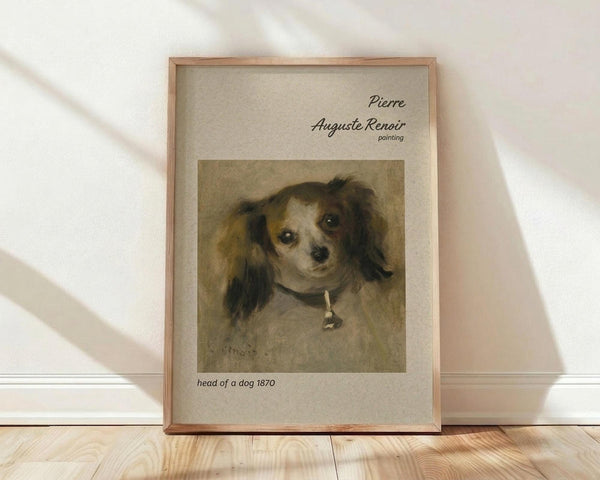 May include: Framed art print leaning against white wall on light wood floor. Print shows a small dog portrait with long dark ears and white face wearing a collar, painted in soft brown and cream tones. Text above reads "Pierre Auguste Renoir painting" and below "head of a dog 1870". Natural wood frame with light colored matting. Soft window light creates shadows on textured white wall.