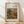 May include: Framed art print featuring Pierre Auguste Renoir's impressionist painting of a Mediterranean farmhouse surrounded by lush green foliage and warm earth tones. The wooden frame sits against a white wall on light wood flooring with soft natural lighting creating diagonal shadows. The artwork shows Renoir's characteristic loose brushwork with vibrant greens, oranges, and blues depicting a rustic building nestled among trees and vegetation.