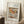 May include: Framed art print leaning against white wall showing Renoir's impressionist still life painting of colorful pears on white fabric. Light wooden frame with white matting displays the soft brushwork and warm tones of red, yellow, and green fruit. Coffee cup on wooden block and textured throw blanket create cozy home setting with natural window light casting shadows.