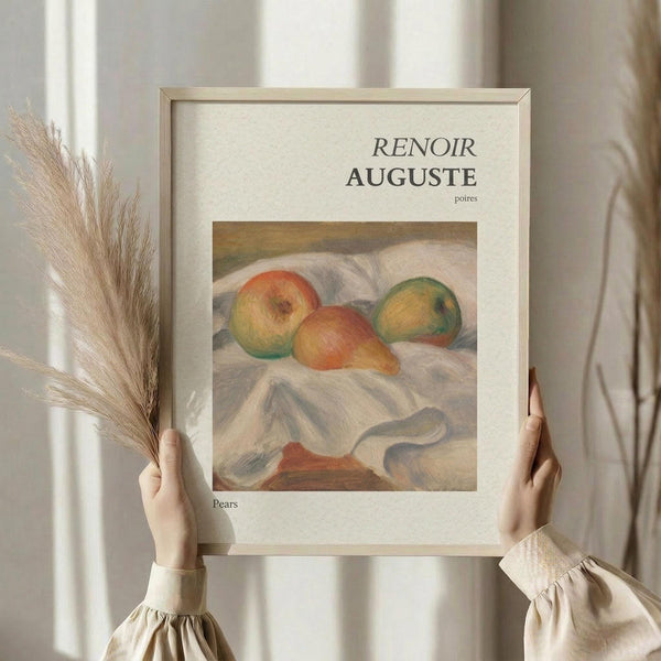 May include: Person in cream ruffled sleeves holding framed art print showing Renoir's impressionist still life painting of pears on white fabric. One hand also holds dried wheat stalks. Soft natural lighting against neutral curtained background. Light wooden frame with white matting displays the classic fruit painting in warm peachy and green tones.