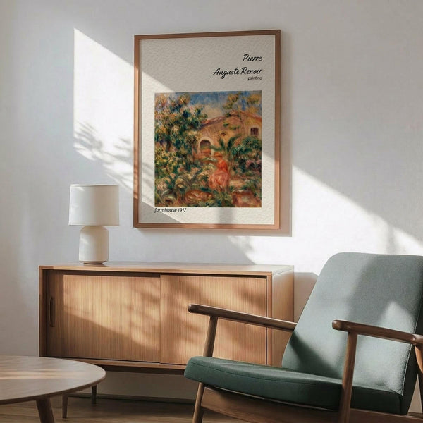 May include: Framed Renoir reproduction print hanging on white wall above wooden sideboard with lamp. Shows colorful Impressionist painting of rural farmhouse scene with lush greenery, warm earth tones, and figure in red. Mid-century modern furniture including sage green upholstered chair with wooden arms and round coffee table. Natural sunlight creates shadows across the minimalist room setting.