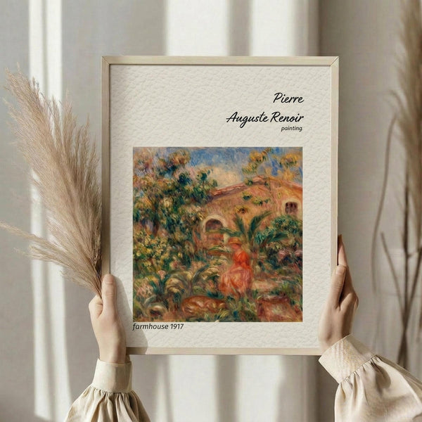 May include: Person in cream sweater holding framed art print featuring Pierre-Auguste Renoir's impressionist farmhouse painting from 1917. Artwork shows warm earth tones with red-tiled roofs, lush greenery, and soft brushstrokes. Light wood frame with cream matting. Dried pampas grass visible to left against neutral curtained background.