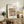 May include: Framed art print displaying Renoir's painting of three pears on white fabric, with "RENOIR AUGUSTE" and "Pears" text on cream mat. Shown leaning against white wall on rustic wooden sideboard alongside fern plant, stacked books, ceramic vases, and brown leather chair visible in foreground of neutral-toned living room setting.