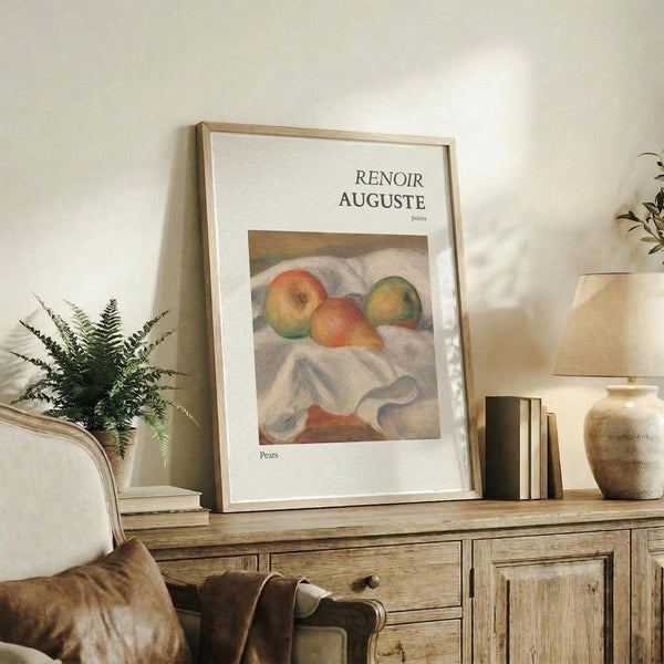 May include: Framed art print displaying Renoir's painting of three pears on white fabric, with "RENOIR AUGUSTE" and "Pears" text on cream mat. Shown leaning against white wall on rustic wooden sideboard alongside fern plant, stacked books, ceramic vases, and brown leather chair visible in foreground of neutral-toned living room setting.