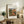 May include: Framed poster print of Renoir's impressionist painting showing a Mediterranean farmhouse with terracotta roof surrounded by lush greenery and foliage in warm earth tones. The artwork is displayed in a thin wood frame on a rustic wooden console table alongside potted plants, books, and ceramic vessels in a neutral-toned living room setting.