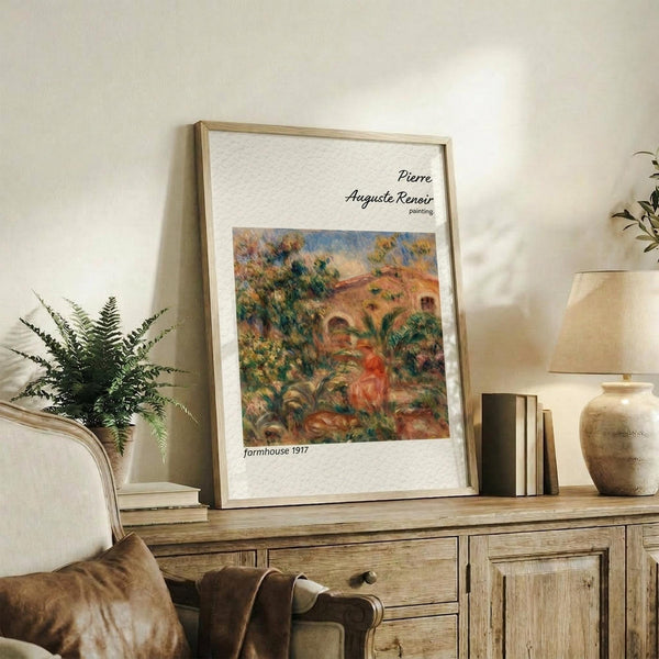May include: Framed poster print of Renoir's impressionist painting showing a Mediterranean farmhouse with terracotta roof surrounded by lush greenery and foliage in warm earth tones. The artwork is displayed in a thin wood frame on a rustic wooden console table alongside potted plants, books, and ceramic vessels in a neutral-toned living room setting.