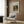 May include: Cream-colored poster featuring a vintage dog portrait painting in warm brown tones, showing a small spaniel-type dog with dark ears and collar. The framed print hangs on wood-paneled wall above a beige upholstered armchair with geometric patterned pillow. Modern brass floor lamp and dark round side table with open book complete the neutral-toned reading nook setting.
