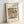 May include: Framed art print leaning against white wall showing Renoir's impressionist still life painting of colorful pears on white fabric. The artwork features soft brushstrokes in warm orange, yellow and green tones. Text reads "RENOIR AUGUSTE" at top and "Pears" at bottom on cream-colored background. Simple light wood frame casts gentle shadow on wall.