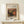 May include: Framed art print featuring Renoir's still life painting of pomegranates and figs in warm orange, yellow and green tones, displayed on a light wood shelf against a cream wall. Small white gift box with evergreen sprig sits beside the natural wood frame.