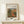 May include: Framed art print displaying Pierre Auguste Renoir's impressionist farmhouse painting from 1917, featuring warm earth tones of orange, green, and brown depicting a Mediterranean villa with arched doorway surrounded by lush foliage. Print has cream matting with black text in thin wooden frame, displayed on light wood console table next to small white wrapped gift with twine and pine sprig against neutral beige wall.