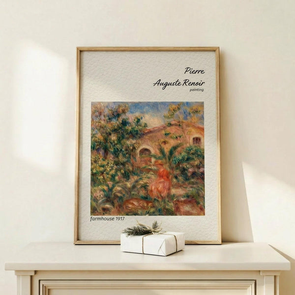 May include: Framed art print displaying Pierre Auguste Renoir's impressionist farmhouse painting from 1917, featuring warm earth tones of orange, green, and brown depicting a Mediterranean villa with arched doorway surrounded by lush foliage. Print has cream matting with black text in thin wooden frame, displayed on light wood console table next to small white wrapped gift with twine and pine sprig against neutral beige wall.