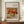 May include: Framed art print featuring Auguste Renoir's 1911 still life painting of red apples, oranges and a green lemon on white fabric. The warm-toned artwork is displayed in a thin brown frame on a wooden console table beside a small white ceramic vase and stacked books against cream wainscoted walls.