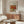 May include: Framed Renoir art print showing colorful still life of apples, oranges and citrus fruits displayed above wooden sideboard in minimalist living room with cream chair, glass coffee table, and modern floor lamp. Warm wood tones complement the vintage French artwork's rich reds, oranges and yellows.