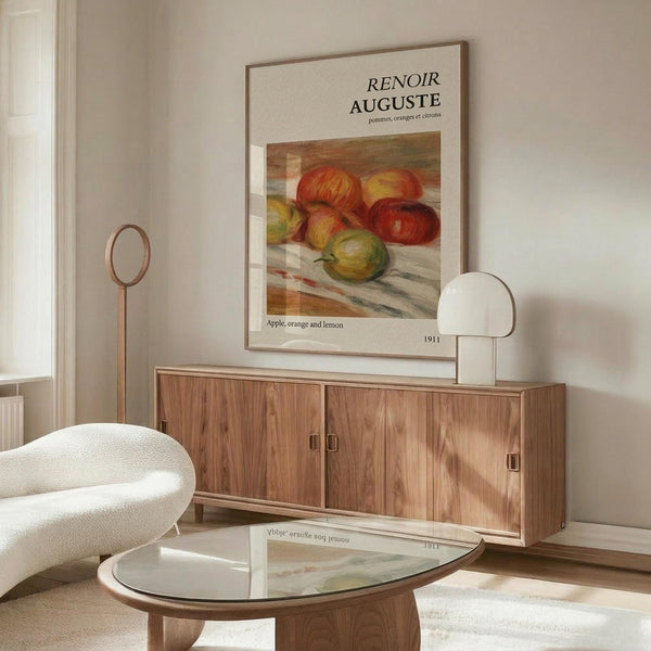 May include: Framed Renoir art print showing colorful still life of apples, oranges and citrus fruits displayed above wooden sideboard in minimalist living room with cream chair, glass coffee table, and modern floor lamp. Warm wood tones complement the vintage French artwork's rich reds, oranges and yellows.