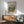 May include: Framed Renoir landscape print hanging on white wall above wooden credenza with white table lamp. Green upholstered mid-century modern armchair with wooden frame sits nearby. Poster shows impressionist painting of countryside scene with chestnut trees, rolling hills, and soft brushstrokes in greens, browns, and blues. Natural sunlight creates shadows across the minimalist room setting.