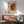 May include: Abstract expressionist style poster with bold red, yellow, black and white paint splatters and dynamic brushstrokes, displayed in wooden frame above mid-century modern credenza. Sage green upholstered armchair with wooden arms sits nearby, white ceramic table lamp on sideboard. Warm sunlight creates geometric shadows on white wall in minimalist living room setting.