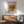 May include: Framed art print of Renoir's fruit still life painting showing red apples, orange, and lemon on white cloth, displayed above wooden sideboard with white ceramic table lamp in modern living room with mint green mid-century armchair and natural sunlight streaming across white walls