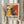 May include: Person in cream sweater holding framed abstract art poster featuring bold red, yellow, black and white paint splatters and dynamic brushstrokes. Wooden frame against neutral wall with dried pampas grass visible to left. Exhibition-style poster design with "POLLOCK" text and dates below colorful chaotic artwork.