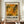 May include: Hands holding a framed abstract art exhibition poster featuring Jackson Pollock-inspired paint splatters in autumn colors of orange, yellow, and green. The poster displays exhibition text and dates, set against cream matting in a brown wooden frame. Scene shows the artwork positioned on a wooden console table with a small cream ceramic vase and books, against white wainscoting wall background.