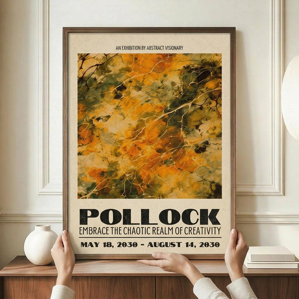 May include: Hands holding a framed abstract art exhibition poster featuring Jackson Pollock-inspired paint splatters in autumn colors of orange, yellow, and green. The poster displays exhibition text and dates, set against cream matting in a brown wooden frame. Scene shows the artwork positioned on a wooden console table with a small cream ceramic vase and books, against white wainscoting wall background.