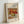 May include: Framed art print featuring Renoir's 1911 still life painting of apples, oranges and a lemon in warm red, yellow and green tones. The poster has white matting with French and English titles, displayed in a light wood frame leaning against a white wall with soft natural lighting.
