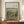 May include: Framed Impressionist landscape painting featuring rolling countryside with chestnut trees, displayed on wooden credenza beside white ceramic vase and stacked books. Warm earth tones of green, brown, and golden hues throughout the pastoral scene. Natural lighting illuminates cream colored walls with white molding in elegant interior setting.